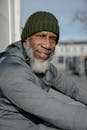 Confident African American bearded male in warm outerwear and hat looking at camera sitting on street in sunny day